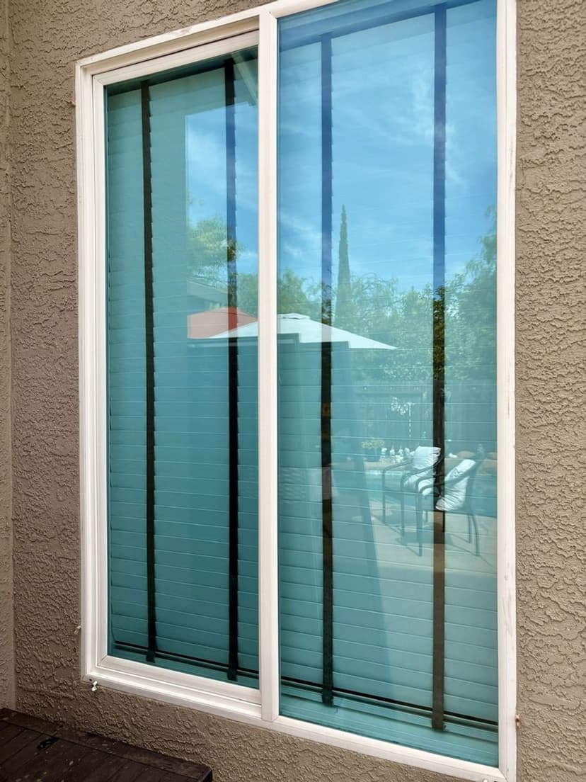 Modern window with light blue blinds, reflecting outdoor patio furniture and greenery.
